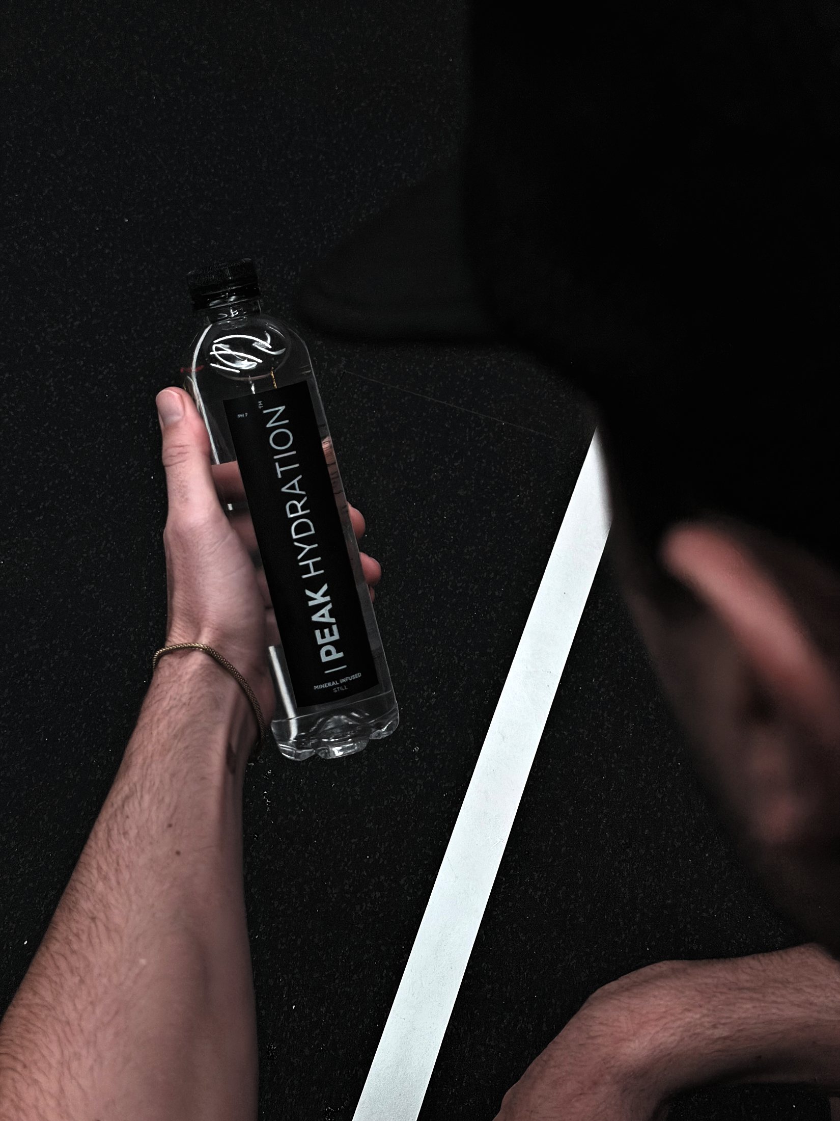 A hand holding a bottle of Peak Hydration still water over a dark training floor